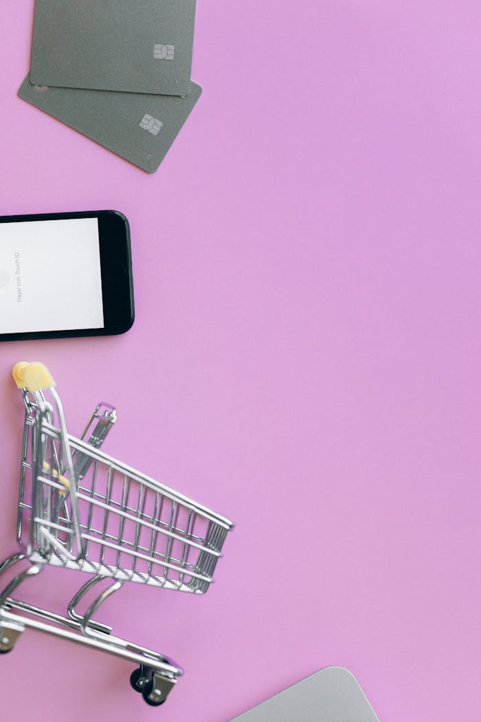 about-01 Top view of miniature shopping cart with credit cards and smartphone on pink background.